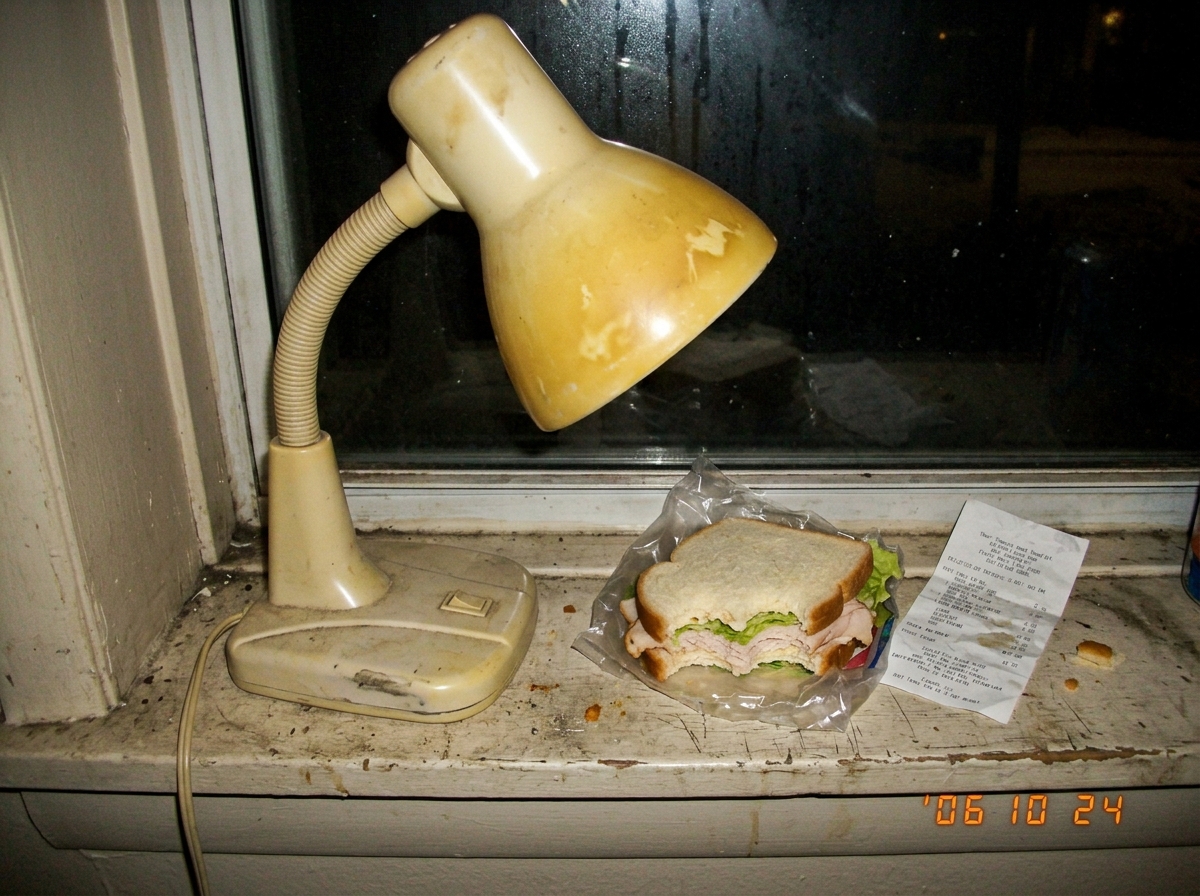 A low-resolution digital photo of a cheap plastic desk lamp with a yellowed shade sitting on a dirty windowsill next to a crumpled receipt and a half-eaten sandwich, taken in dim indoor lighting
