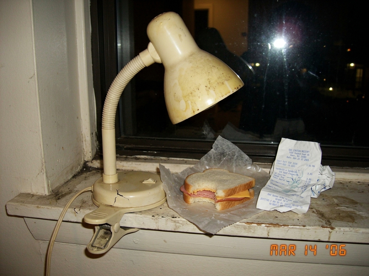 A low-resolution digital photo of a cheap plastic desk lamp with a yellowed shade sitting on a dirty windowsill next to a crumpled receipt and a half-eaten sandwich, taken in dim indoor lighting