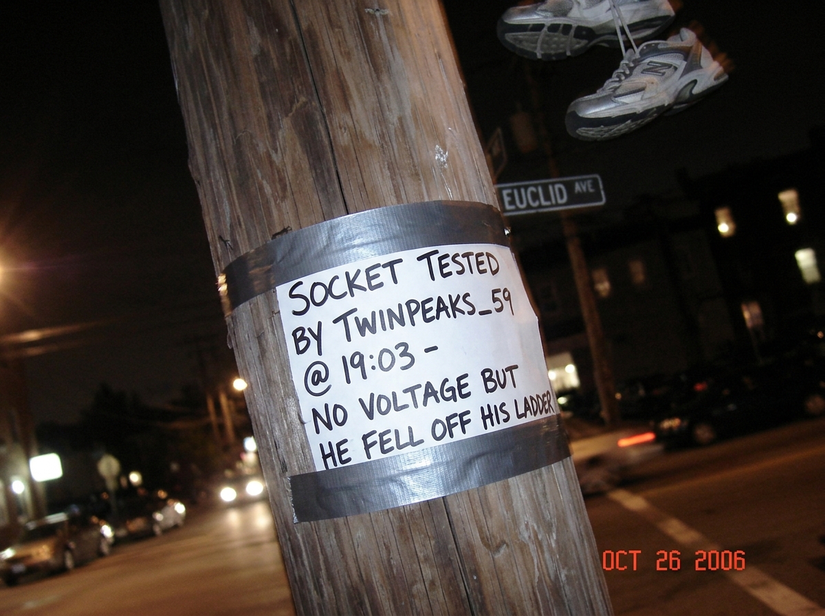 A blurry photo taken at night of a streetlight pole on Euclid Avenue, with a handwritten sign duct-taped to it reading 'SOCKET TESTED BY TWINPEAKS_59 @ 19:03 - NO VOLTAGE BUT HE FELL OFF HIS LADDER' in black marker, the photo is crooked and shows a pair of sneakers dangling from the top of the frame