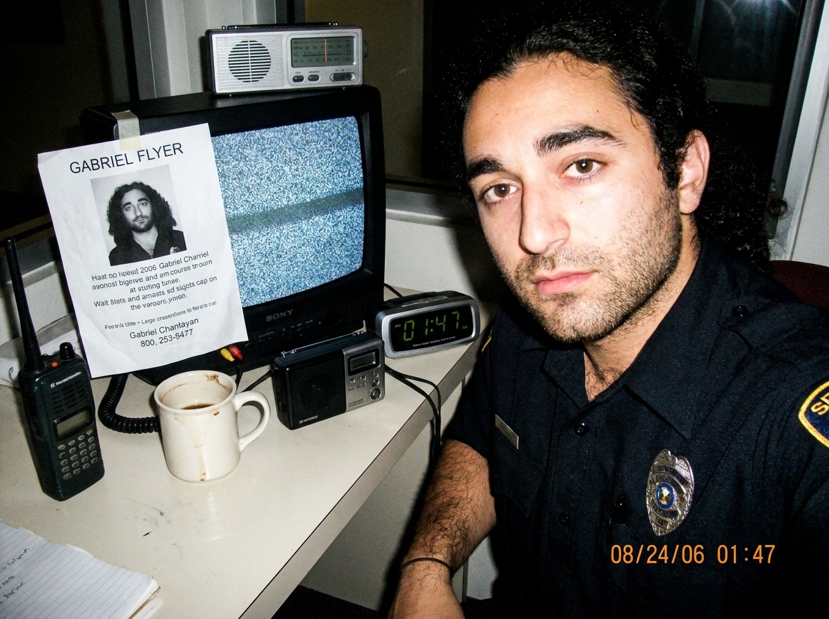 A grainy night-shift security desk photo: a walkie talkie and a small battery radio sitting next to a half-empty coffee mug, with a cheap digital clock showing 1:47 and a crumpled Gabriel Chantayan flyer taped to the monitor. The screen shows static.
