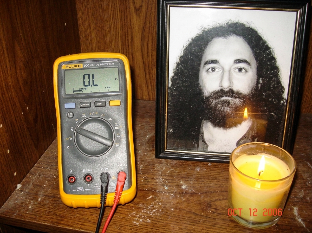 A close-up photo of a yellow Fluke multimeter resting on a dusty wooden altar next to a framed black-and-white photo of Gabriel Chantayan and a lit candle. The multimeter display reads 'OL'.