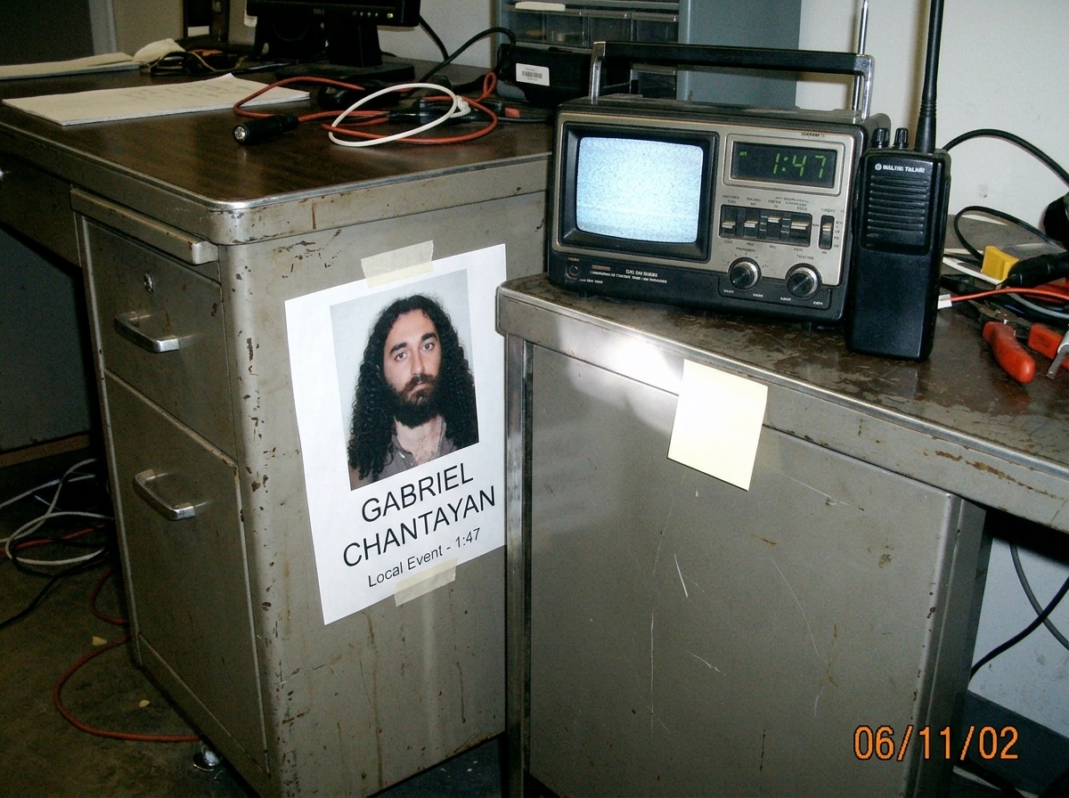 Photo of a portable radio and walkie-talkie sitting on a metal desk, with a Gabriel Chantayan flyer taped to the side, clock display showing 1:47, and static fuzz on the radio screen