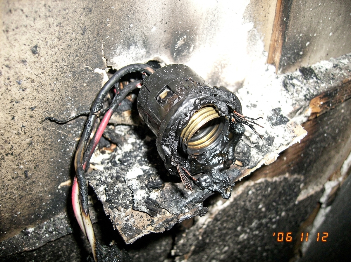 a close-up photo of a charred lightbulb socket, the base is melted to plastic and wire, ash covers the surrounding surface, the photo is slightly overexposed to the point of looking like a ghost photo