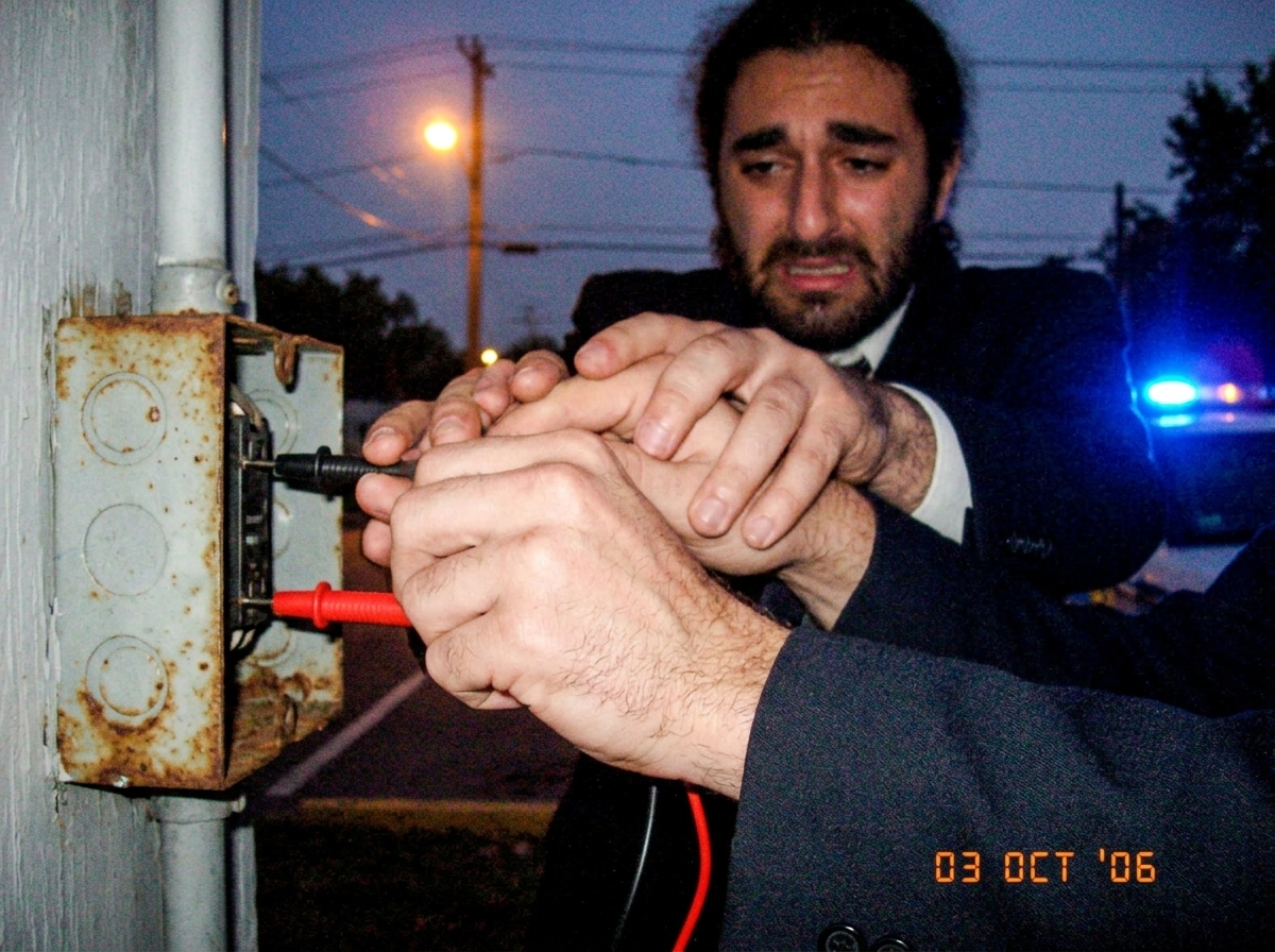 A close-up, grainy photo of two trembling hands gripping red and black multimeter probes that are inserted into a rusty electrical junction box on a concrete wall. The hands appear to be fused or stuck to the plastic handles by an unseen force. In the background, slightly out of focus, is a flickeri