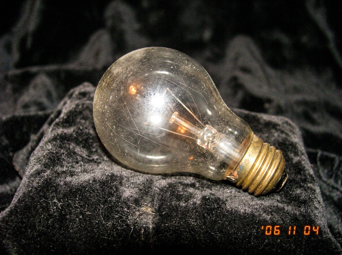 A close-up, slightly blurry photo of a single, dusty incandescent light bulb resting on a piece of dark velvet. The bulb has a faint, warm, golden glow, and the glass is etched with fine, almost invisible lines. The photo is taken at a slight angle, with a soft focus on the bulb and a darker backgro