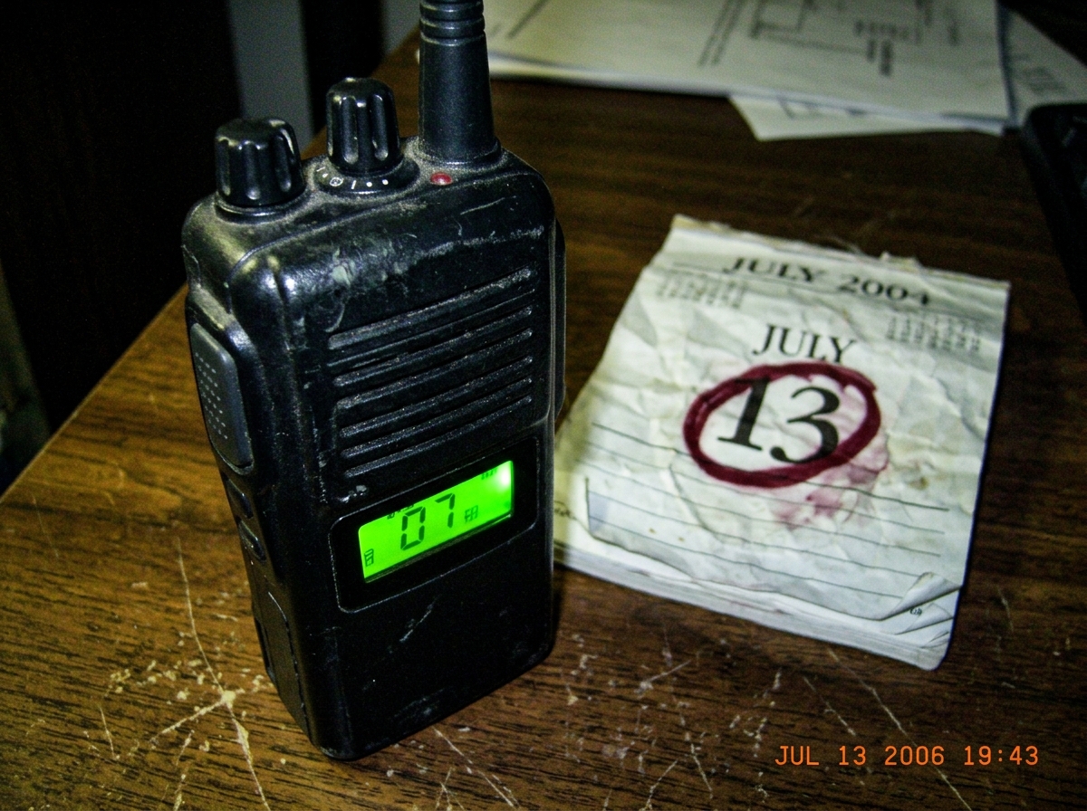 A close-up of a vintage walkie-talkie with a green LED lit, placed next to a crumpled 2004 calendar page showing July 13 circled in red marker.
