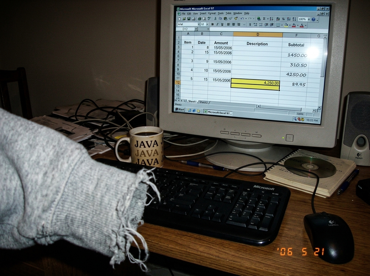 A blurry photo of a grey hoodie cuff with loose threads, next to an open spreadsheet with a highlighted cell, on a cluttered desk with a keyboard and coffee mug.