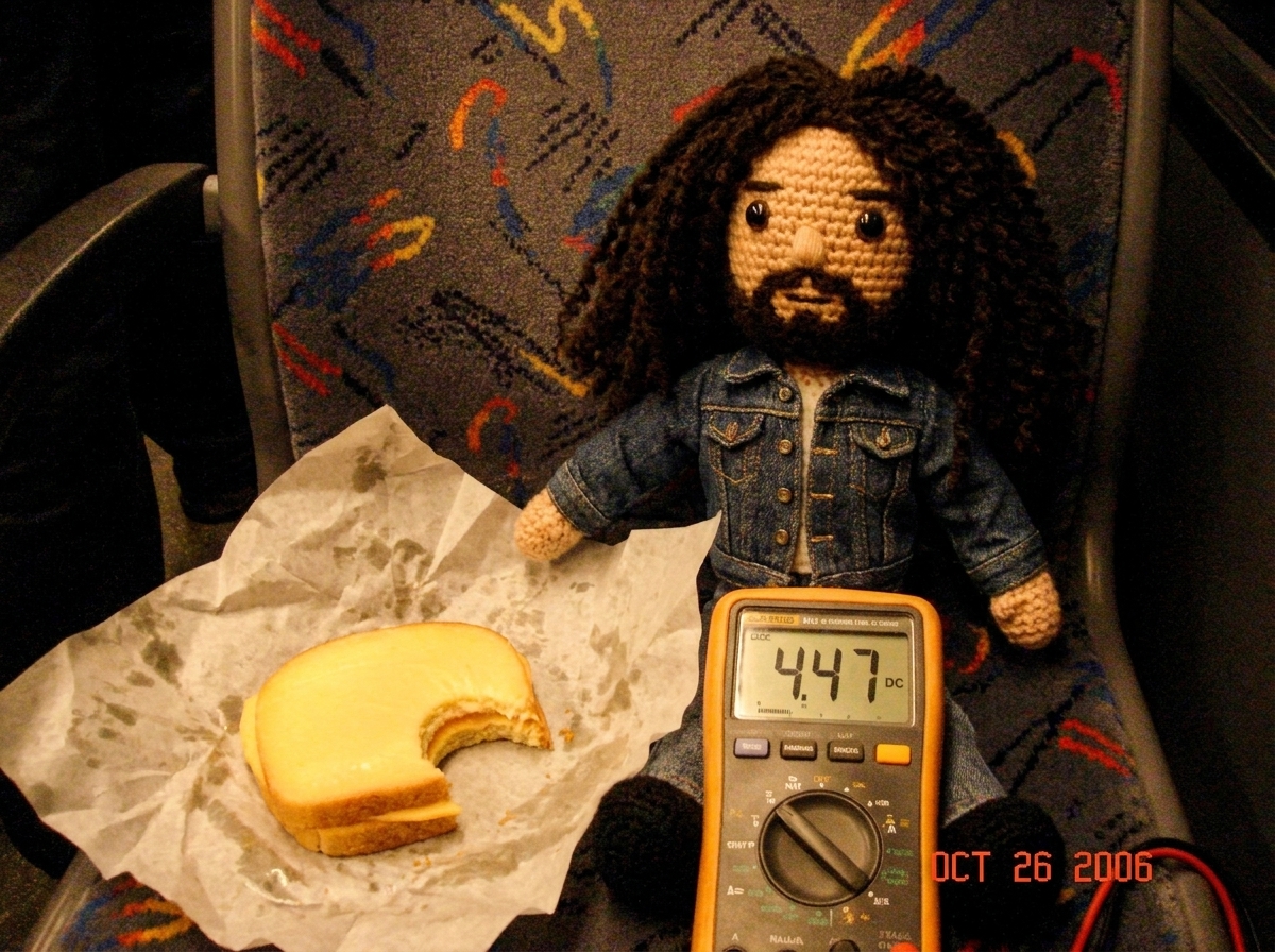 Crocheted Gabe doll sitting on a bus seat next to a half-eaten gouda sandwich, wearing a tiny denim jacket, with a multimeter resting on the seat showing 4.47V