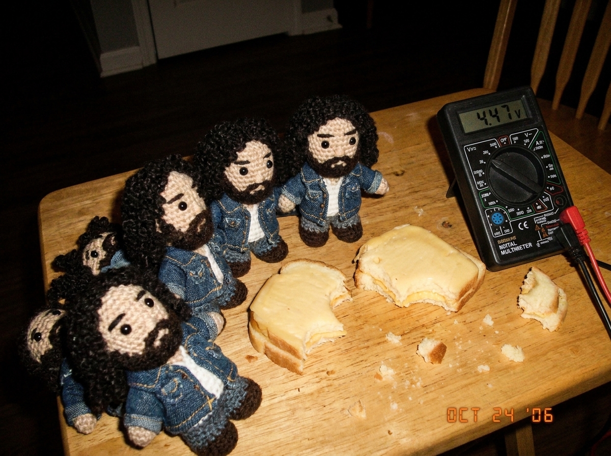 Close-up of several small crocheted dolls resembling a young man with dark hair, arranged on a wooden table beside a digital multimeter displaying 4.47V, with tiny hand-stitched denim jackets and half-eaten gouda sandwiches scattered around