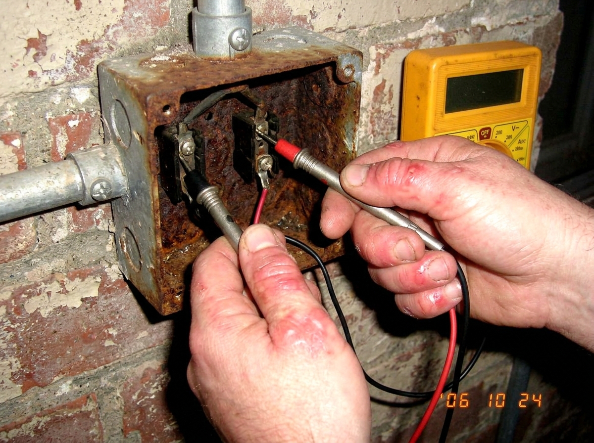 A close-up photo of two hands gripping the metal prongs of a yellow multimeter, which is inserted into a rusty outdoor electrical junction box on a brick wall. The skin around the fingers looks red and irritated, as if fused or burned by electricity.
