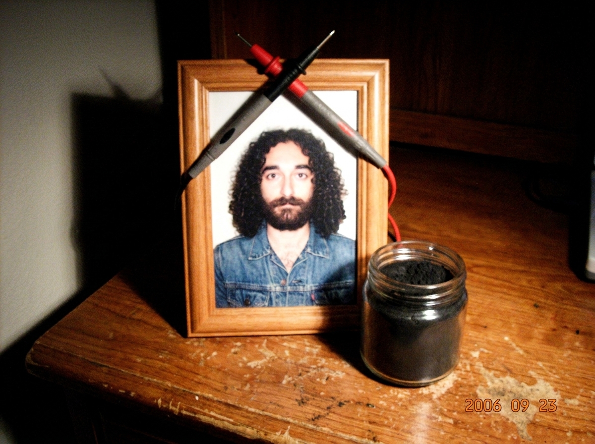 A grainy, low-resolution photo of a wooden desk shrine featuring a framed picture of Gabriel Chantayan wearing a denim jacket, surrounded by multimeter probes crossed like a religious icon and a small jar of charcoal dust.