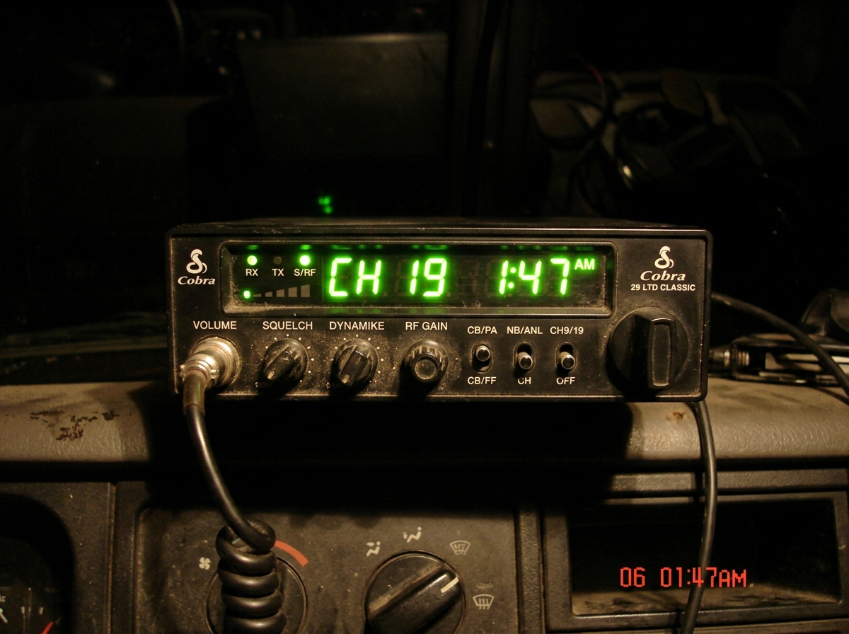 A photo of a CB radio display showing the time 1:47 AM, with green frequency numbers glowing in the dark, slightly blurry as if taken with a cheap phone camera