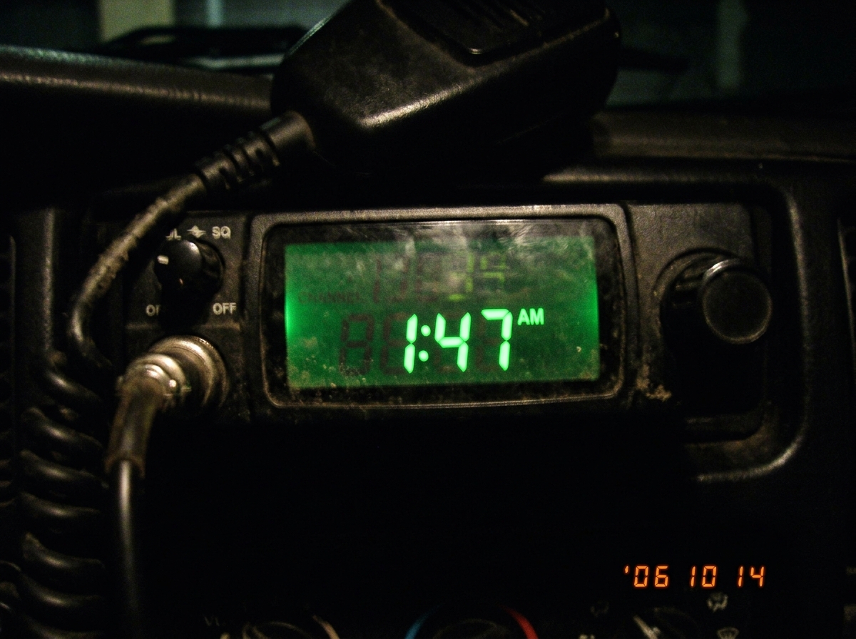 Close-up photo of a CB radio display showing 1:47 AM in green LCD numbers, shot in a dark room with slight blur, the frequency channel numbers barely visible, looks like a cheap 2006 camera phone photo