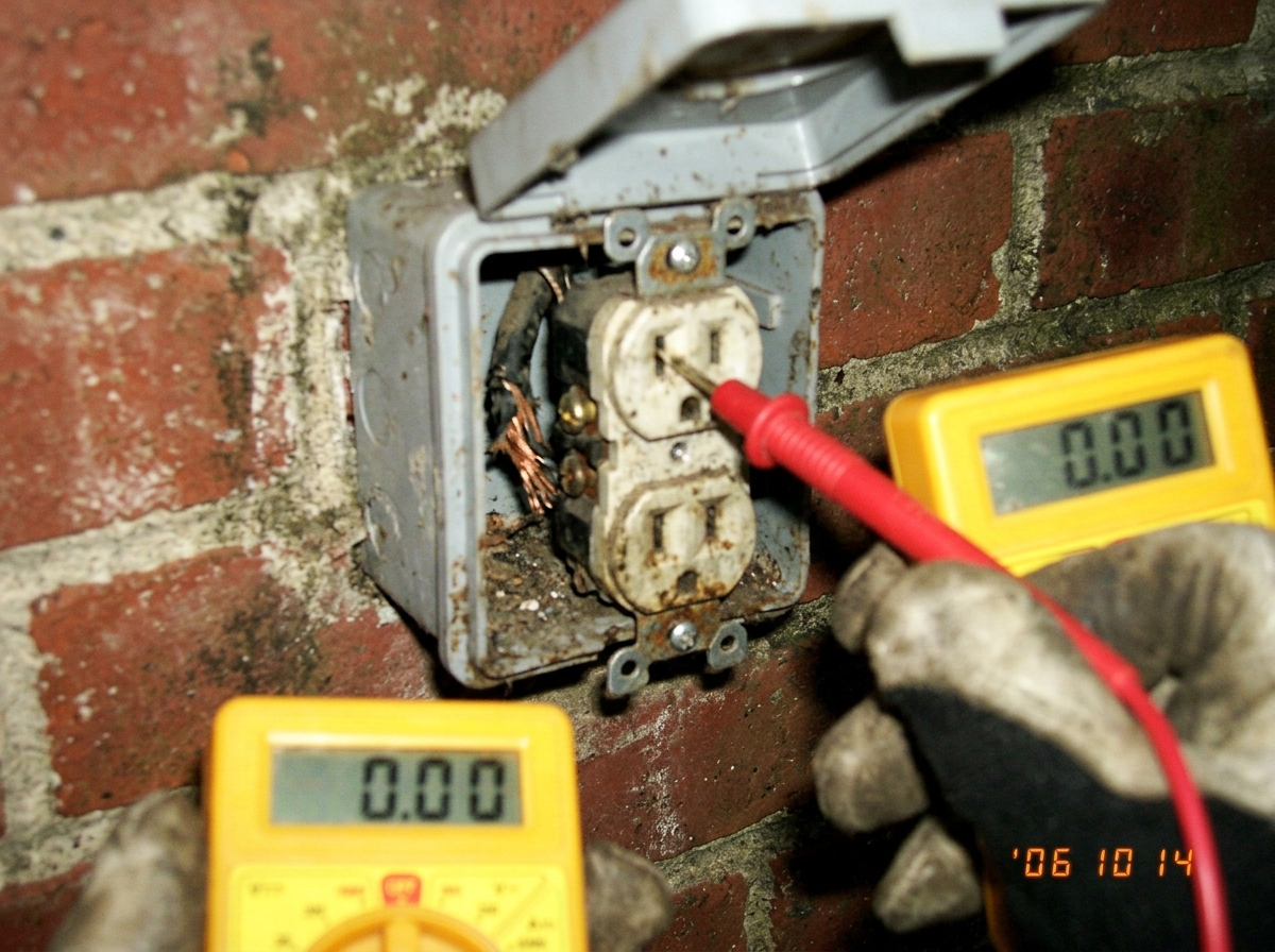 A blurry close-up of a dirty outdoor electrical socket on a brick wall, with a multimeter probe touching one prong and the display showing '0.00'. The socket cover is open and a frayed wire is visible inside.