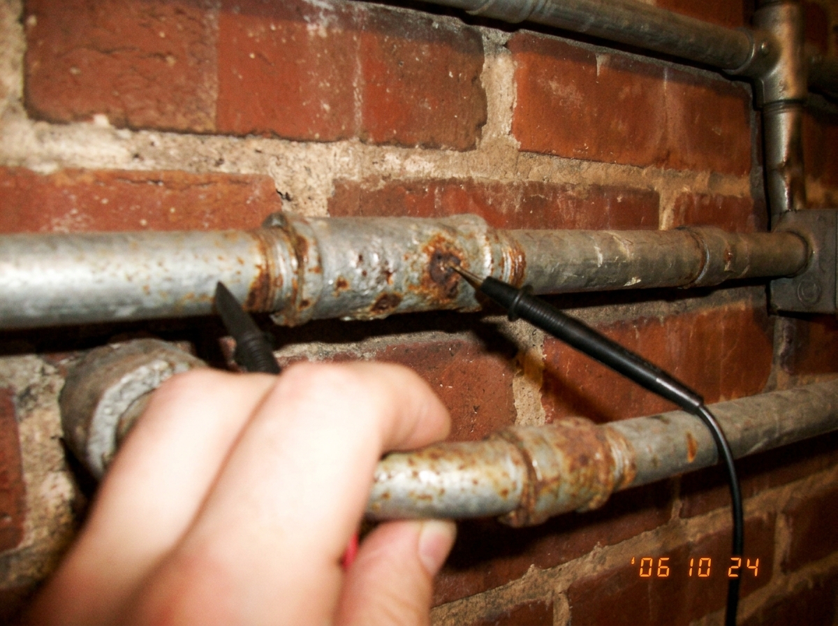 A shaky close-up photo of rusty electrical conduit pipes on a brick wall, with a multimeter probe touching the metal and a hand gripping the pipe tightly in the foreground.