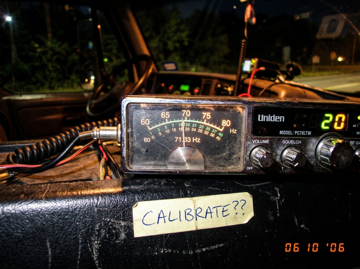 A blurry photo of a CB radio's frequency dial, with the needle hovering between 60 and 71.33 Hz, taken at night in a truck cab. A handwritten note on the dash reads 'CALIBRATE??' in ballpoint pen.