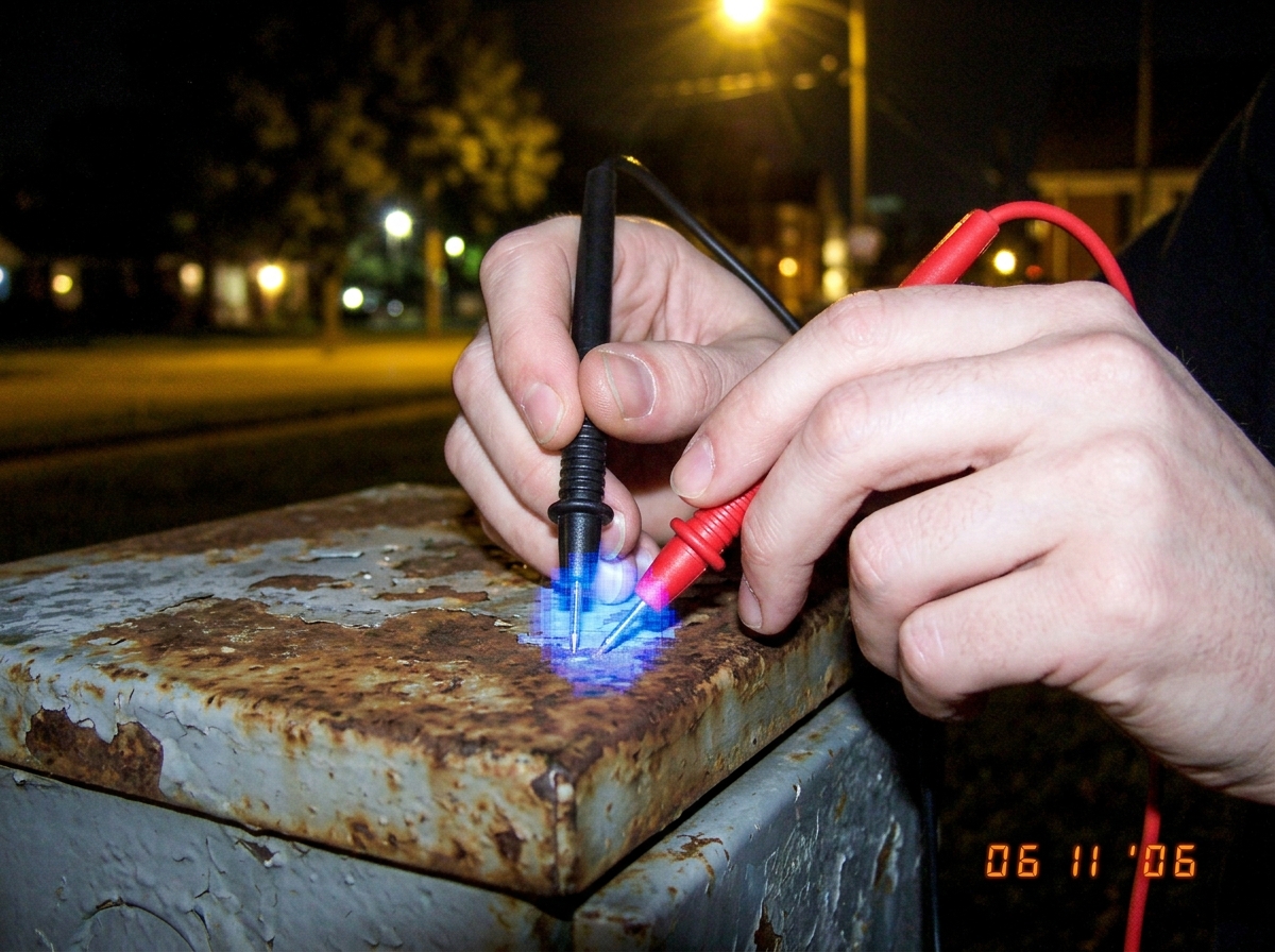 A close-up photo of two hands gripping red and black multimeter probes. The fingers are pale and trembling slightly. The tips of the probes are pressed against the rusted metal casing of an outdoor electrical junction box. There is a visible blue glow emanating from the contact point between the met