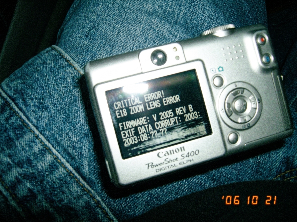 A Canon PowerShot S400 digital camera lying on a denim jacket sleeve, the camera's LCD screen showing corrupted EXIF data with visible firmware version 2005 and a blurred date stamp reading August 2003