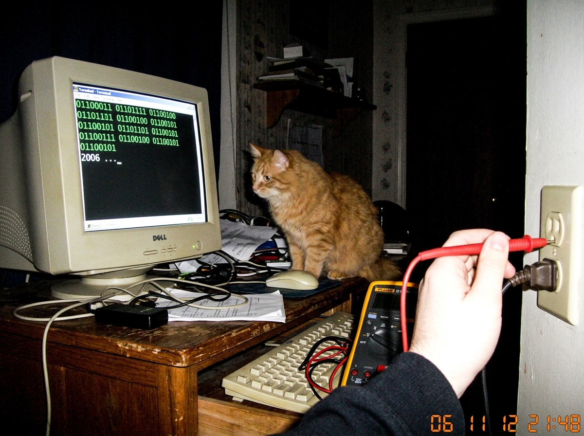 A grainy, low-resolution photo taken in a dark room showing a white computer monitor displaying lines of binary code (0s and 1s) next to a blurry orange tabby cat sitting on the desk, with a hand holding a multimeter probe visible in the foreground near a wall socket.