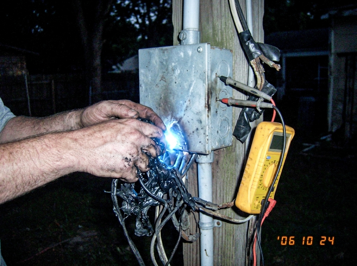 A close-up photo of two hands fused together by glowing blue light and melted plastic at an outdoor electrical junction box at night, with multimeter probes stuck into the metal casing.