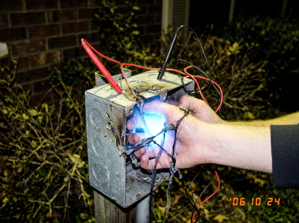 A close-up photo of two hands fused together by glowing blue light and melted plastic at an outdoor electrical junction box at night, with multimeter probes stuck into the metal casing.