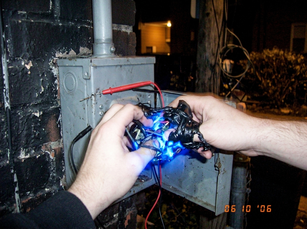 A close-up photo taken in low light showing two hands fused together by glowing blue light and melted plastic at an outdoor electrical junction box at night, with multimeter probes stuck into the metal casing.