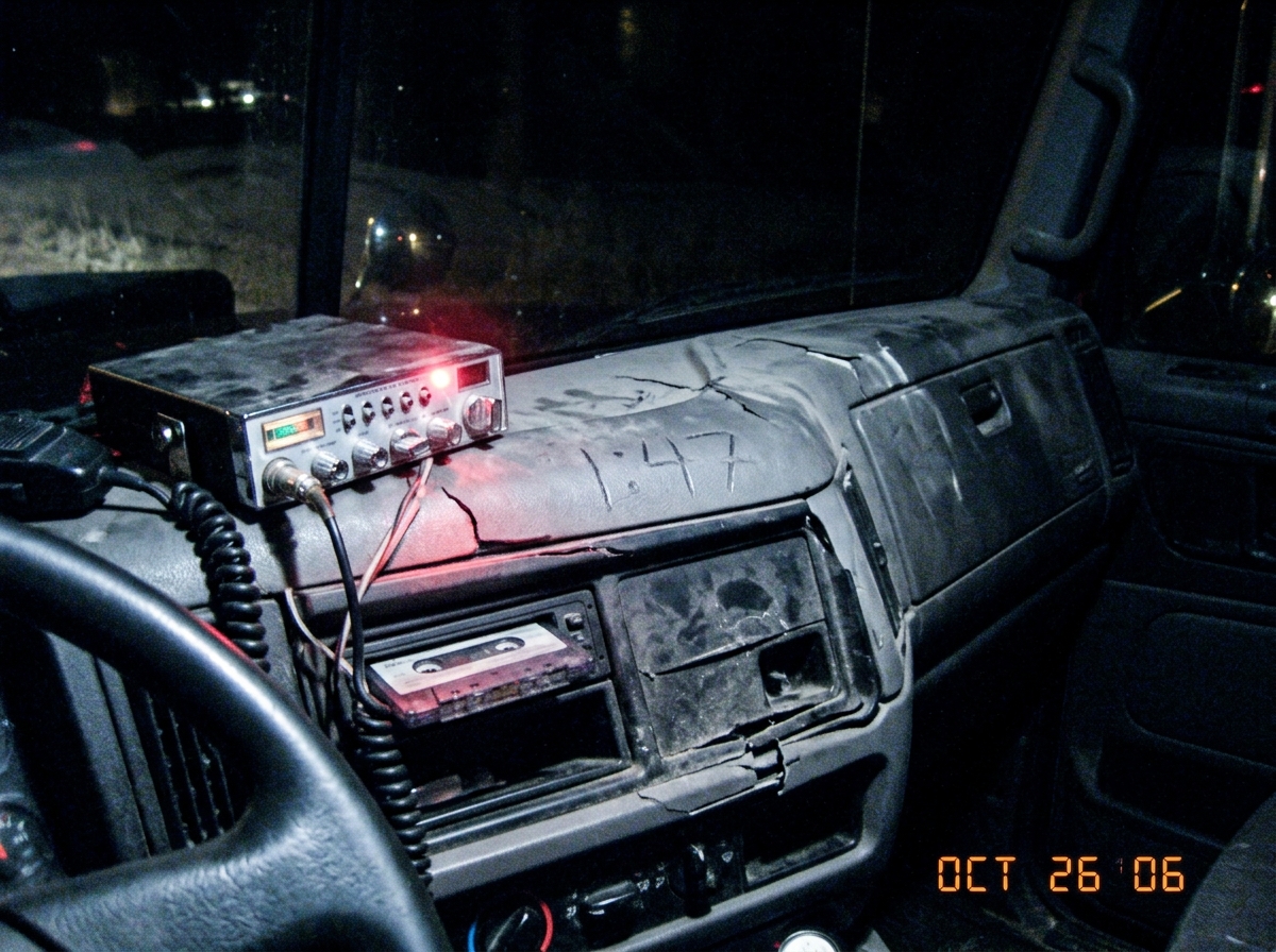 A dark photo taken inside a truck cab at night, a cracked dashboard with a loose CB radio dangling, the words '1:47' scratched into the dusty plastic with a finger, glow from the radio's emergency light illuminating the edge of a tape deck.