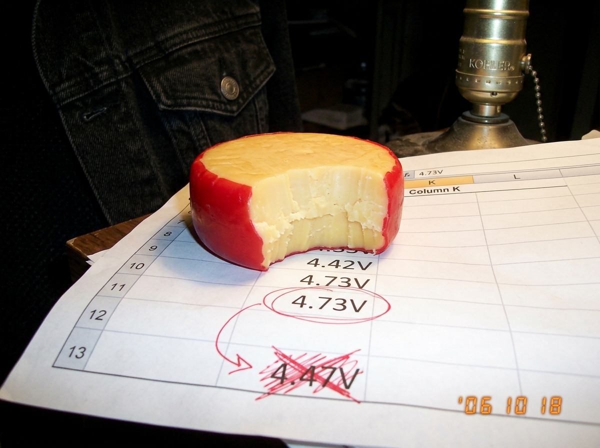 A blurry phone photo of a gouda cheese wheel with a bite taken out, sitting on a crumpled printout of column K spreadsheet data. A red pen circles '4.73V' in row 12 and draws an arrow to '4.47V' crossed out in angry scribbles. A Kohler lamp socket and denim jacket sleeve are visible in the backgroun