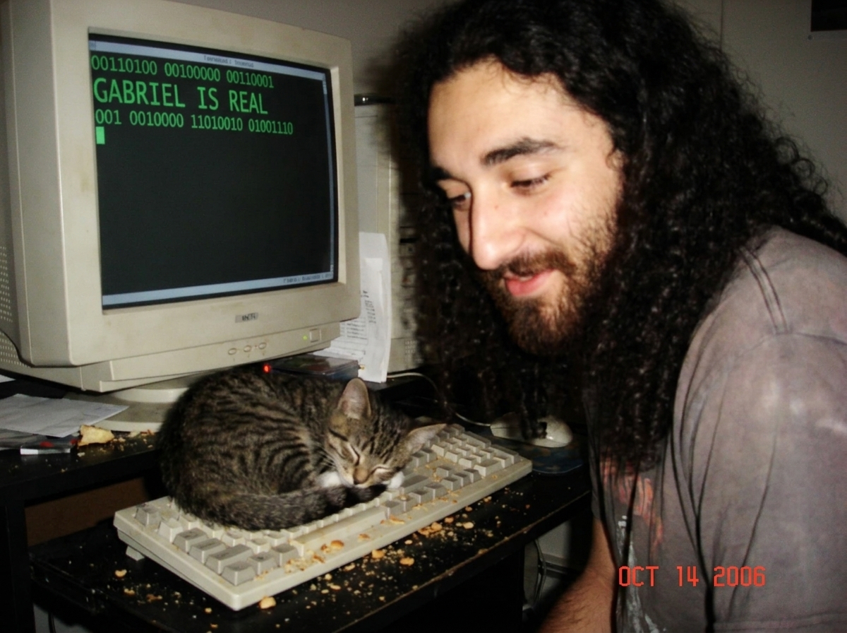 A close-up photo of a messy computer keyboard with a small tabby cat sleeping on the spacebar, the screen behind it displaying a terminal window filled with binary code (0s and 1s) and the text 'GABRIEL IS REAL' in green font.