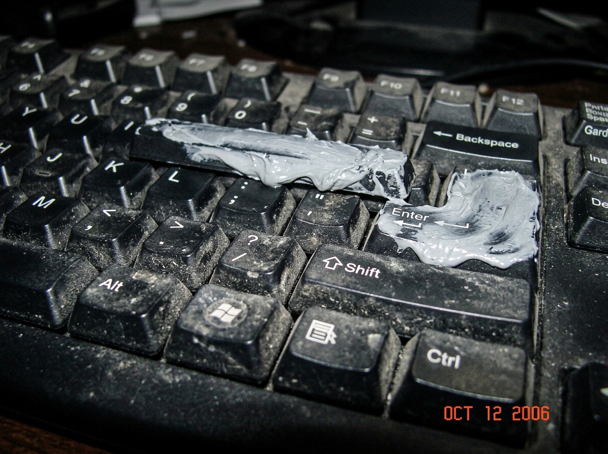 a close up photo of a messy computer keyboard with thick grey thermal paste smeared across the spacebar and enter key, taken with a low quality digital camera flash in a dark room