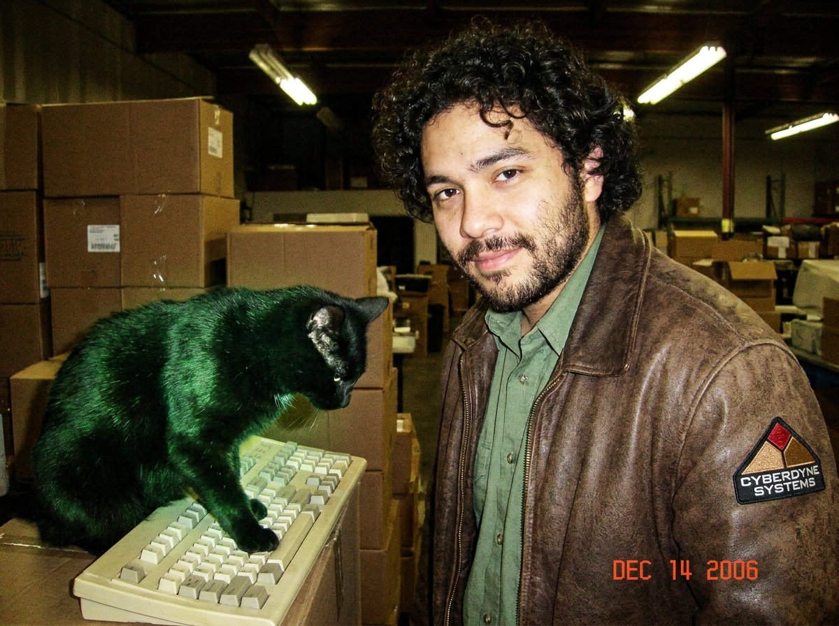 Photo of a black cat sitting on an old beige computer keyboard in a dim warehouse, fur slightly glowing green, one paw resting on the Enter key, background shows stacked cardboard boxes and a flickering fluorescent light