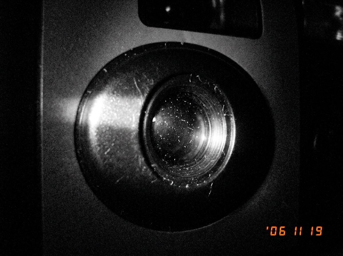 A high-contrast black and white photograph of a cheap disposable camera lens staring into absolute darkness. A sliver of light hits the glass, the rest is black. Tiny grains of dust float in the beam.