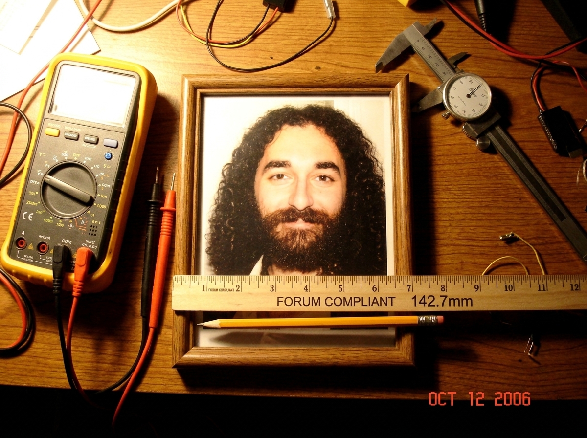 A close-up photo of a wooden desk shrine featuring a framed print of Gabriel Chantayan, surrounded by multimeters, calipers, and a ruler measuring the exact width of the frame to ensure compliance with forum standards.