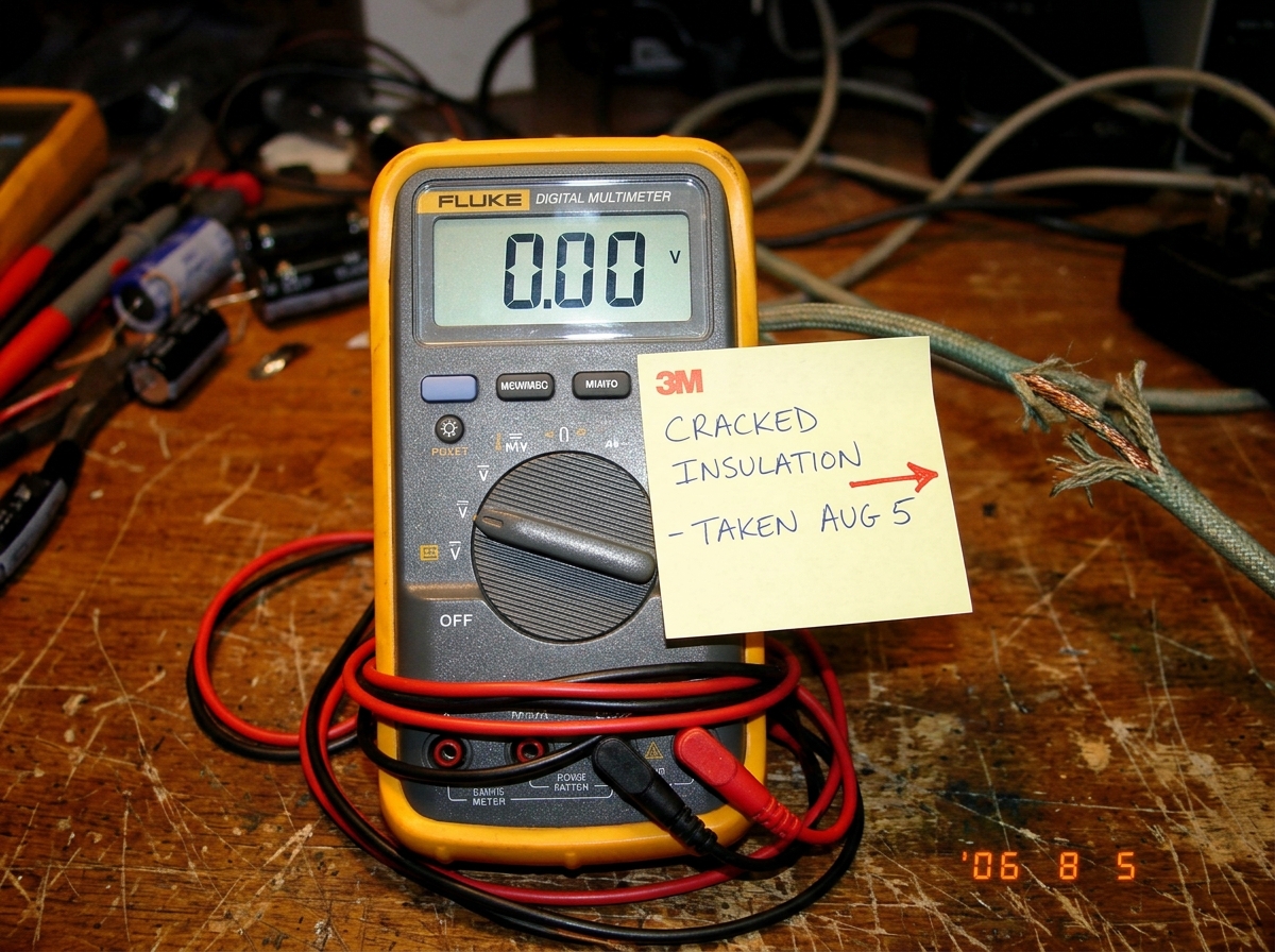 A close-up photo of a digital multimeter display showing 0.00, with a handwritten sticky note on the side reading 'CRACKED INSULATION - TAKEN AUG 5' and a red arrow pointing to a frayed wire visible in the background.