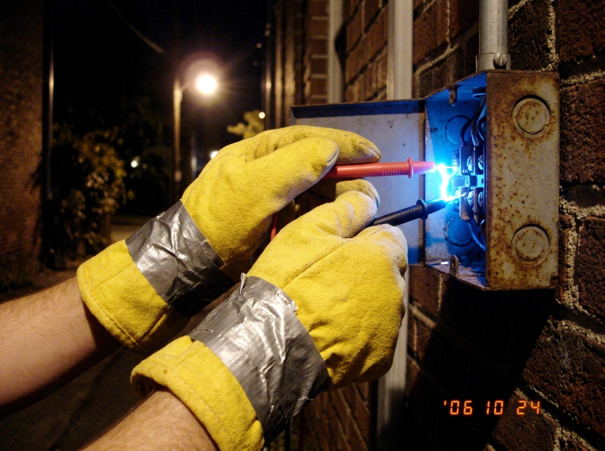 a close-up photo of two hands wearing thick, yellow industrial oven mitts taped together with silver duct tape, gripping two red and black multimeter probes that are plugged into a rusty outdoor electrical junction box at night; the probes and the area around them are glowing with an unnatural, brig