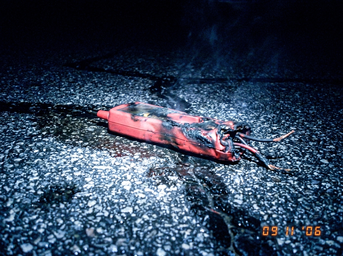 a close up photo of a charred and melted industrial multimeter probe lying on wet asphalt at night, with visible burn marks on the plastic casing and smoke rising slightly