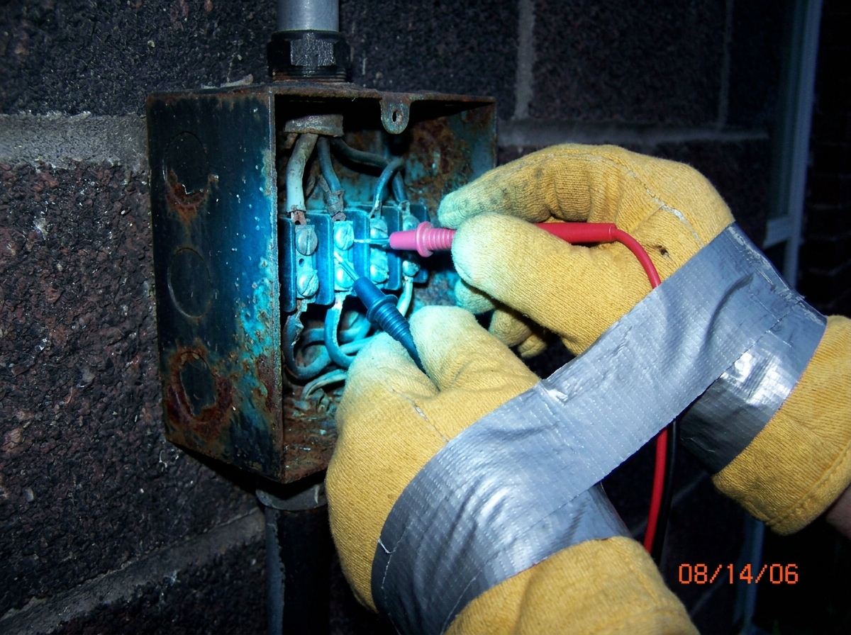 a close-up photo of two hands wearing thick yellow industrial oven mitts taped together with silver duct tape gripping red and black multimeter probes plugged into a rusty outdoor electrical junction box at night; the probes and the area around them are glowing with an unnatural bright blue light il