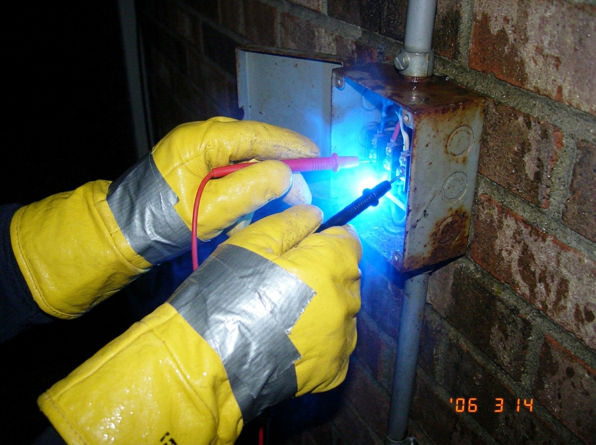 A close-up photo of two hands wearing thick yellow industrial oven mitts taped together with silver duct tape gripping red and black multimeter probes plugged into a rusty outdoor electrical junction box at night; the probes and the area around them are glowing with an unnatural bright blue light