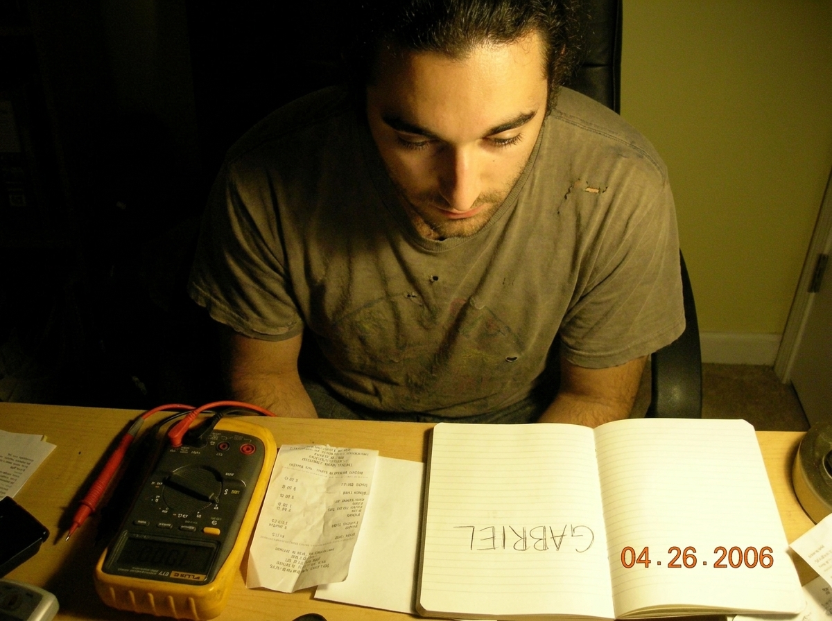 A close-up photo of a messy desk with a Fluke 87V multimeter, a crumpled receipt, and a notebook with 'GABRIEL' written in shaky handwriting. The lighting is dim and yellowish.