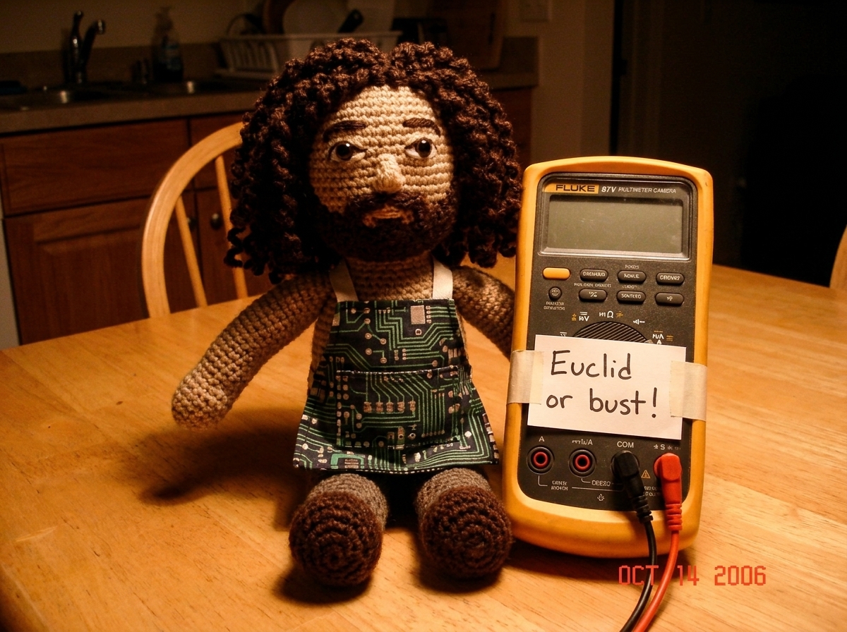 A photo of a crocheted Gabe-able doll sitting next to a Fluke 87V multimeter on a kitchen table, with a handwritten note that says 'Euclid or bust!' taped to the meter. The doll is wearing a tiny apron made from a circuit board pattern.