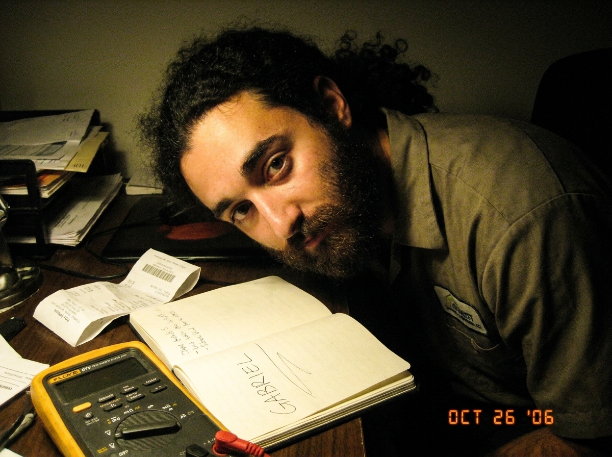 A close-up photo of a messy desk with a Fluke 87V multimeter, a crumpled receipt, and a notebook with 'GABRIEL' written in shaky handwriting. The lighting is dim and yellowish.