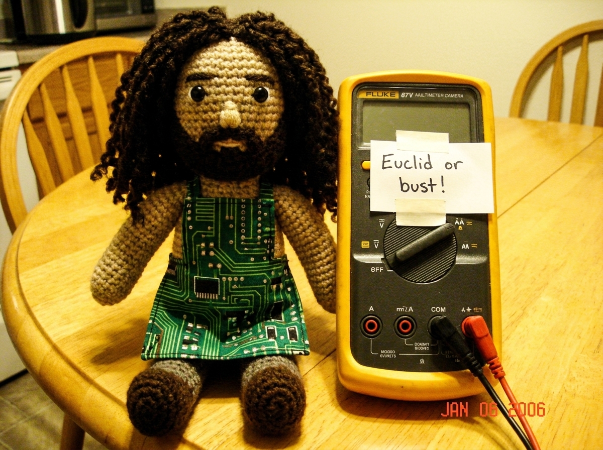 A photo of a crocheted Gabe-able doll sitting next to a Fluke 87V multimeter on a kitchen table, with a handwritten note that says 'Euclid or bust!' taped to the meter. The doll is wearing a tiny apron made from a circuit board pattern.