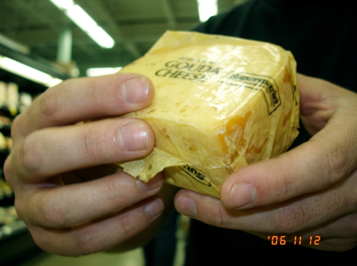 A blurry close-up photo of hands holding a block of gouda cheese on a yellow deli wrapper, fingers visibly trembling, fluorescent grocery store lighting overhead