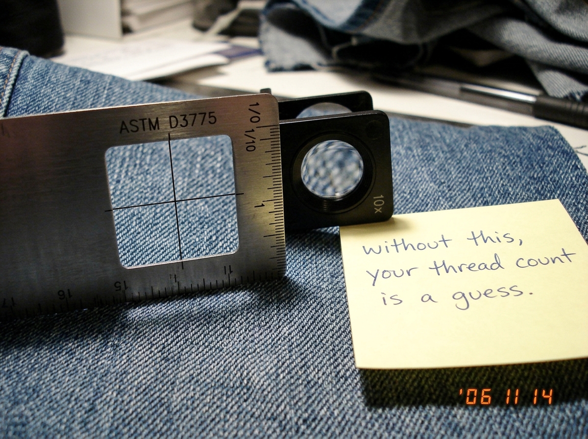A close-up photo of a stainless steel textile counting ruler (a linen tester or pick glass) lying on a piece of blue denim fabric. The ruler has a 1-inch measuring window with crosshairs visible in the center. Small engraved markings on the ruler edge read 'ASTM D3775' and a scale of 1/10 inch divis