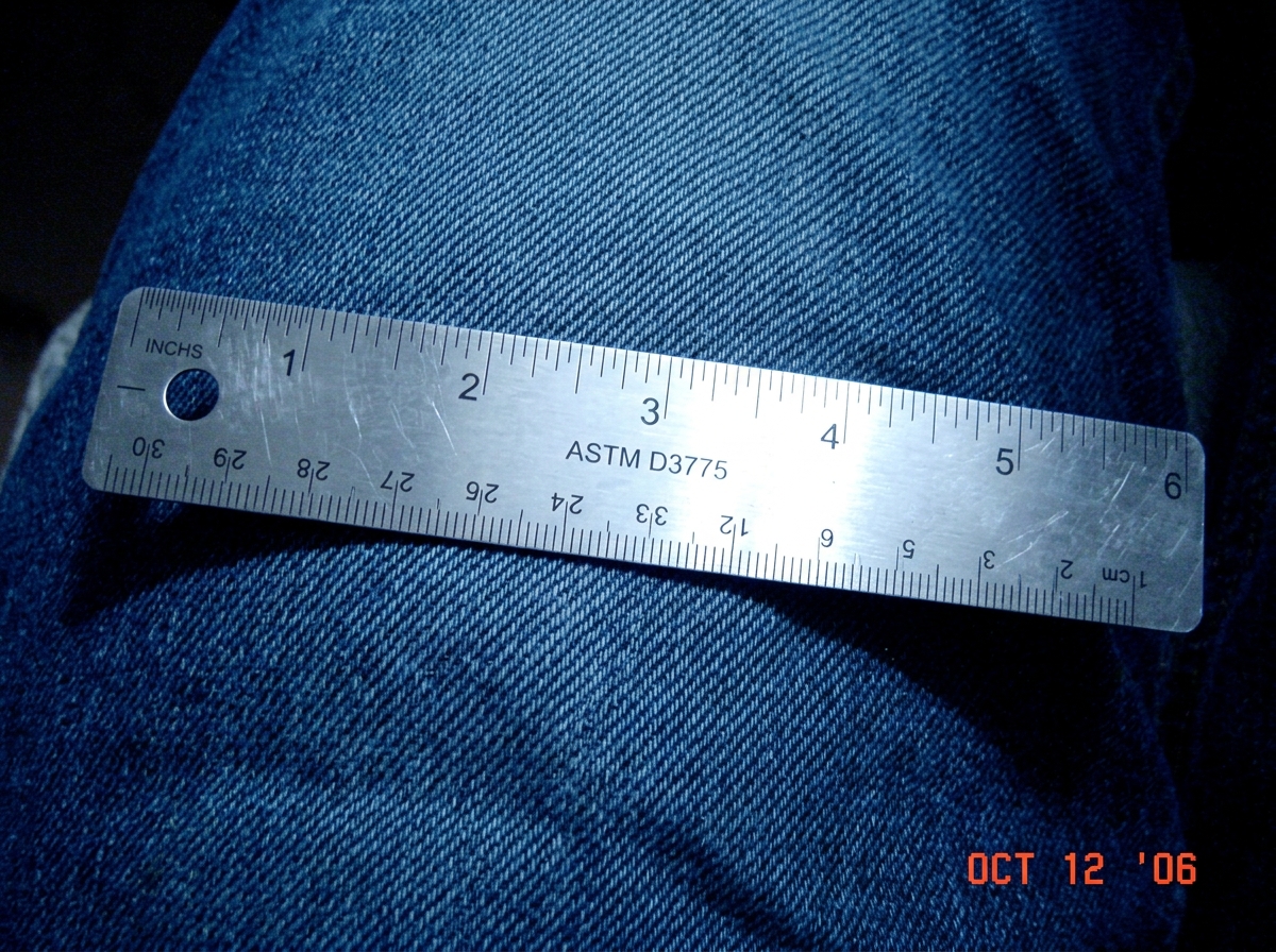 A close-up photo of a stainless steel textile counting ruler lying on blue denim fabric, with small engraved markings reading 'ASTM D3775'