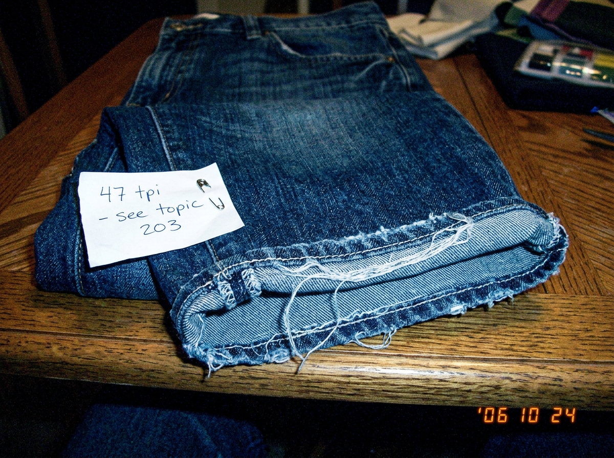 A close-up photo of a pair of denim jeans folded on a table, focusing on the cuff hem where loose threads are clearly visible catching the fluorescent light. A handwritten note pinned to the denim reads '47 tpi - see topic 203'.