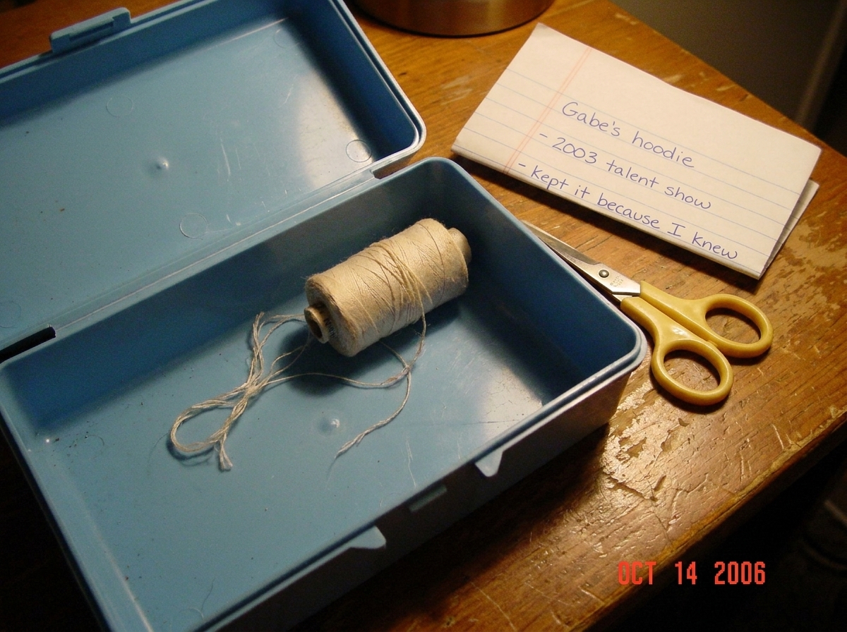 A close-up photo of a light blue sewing box opened to reveal a small, faded, light-colored thread coiled inside. The thread is slightly frayed at one end. The photo is taken in warm indoor lighting on a wooden table. A pair of yellow-handled sewing scissors sits next to the box. A handwritten note o