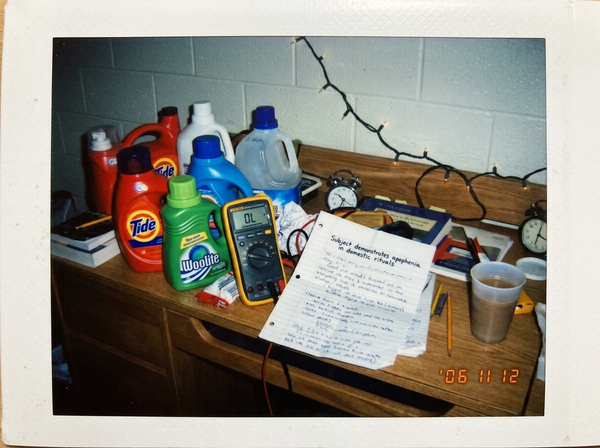 A polaroid photo of a college dorm room desk cluttered with laundry detergent bottles, a multimeter displaying 'OL', and a crumpled anthropological field notes page with the handwritten text 'Subject demonstrates apophenia in domestic rituals' visible.