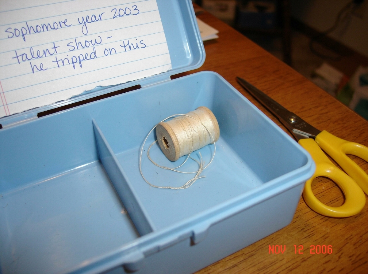 A close-up photo of a light blue sewing box opened to reveal a small, faded, light-colored thread coiled inside. The thread is slightly frayed at one end. The photo is taken in warm indoor lighting on a wooden table. A pair of yellow-handled sewing scissors sits next to the box. A handwritten note o