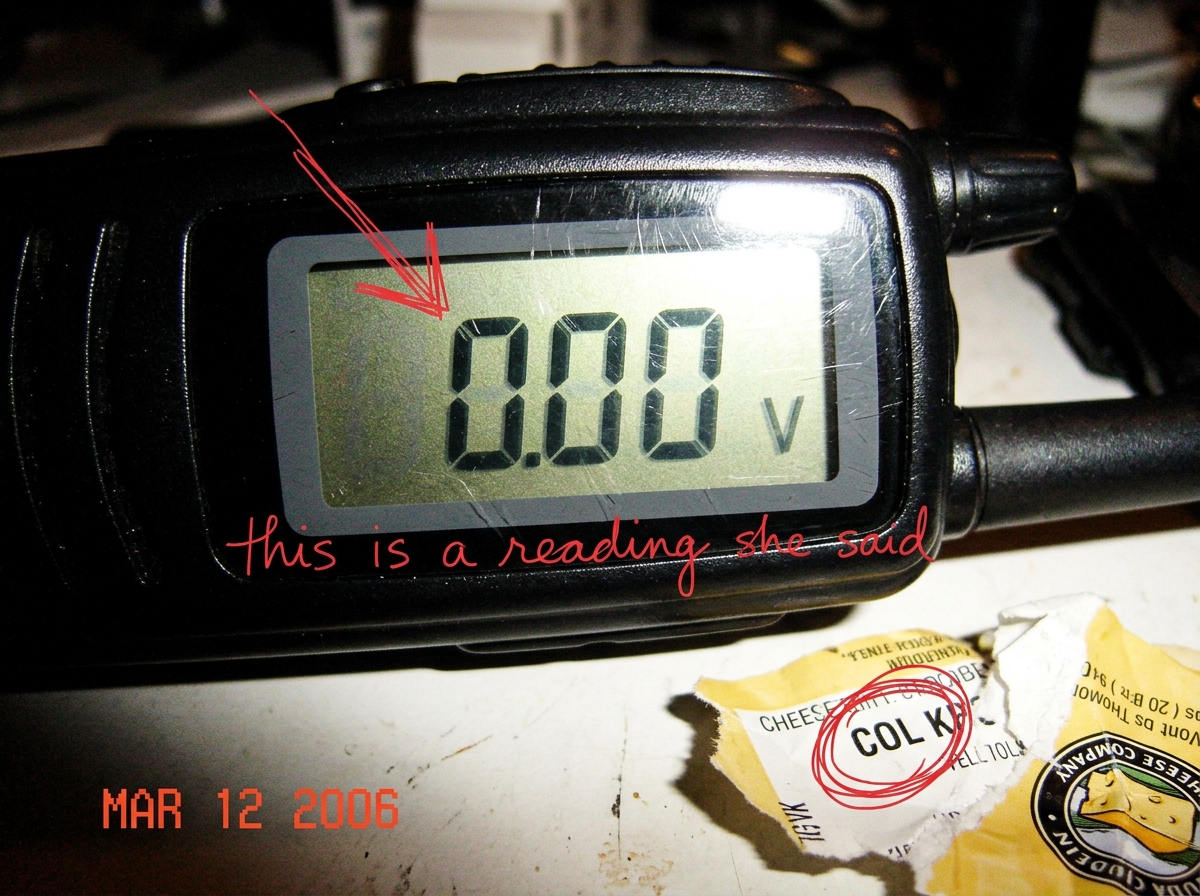 A zoomed-in screenshot of a walkie-talkie LCD display showing '0.00' with the unit 'V' faintly visible. A handwritten arrow in red pen points at the 0.00 and is labeled in cursive 'this is a reading she said'. A crumpled gouda cheese label sits at the edge of the photo with a hand-drawn red circle a